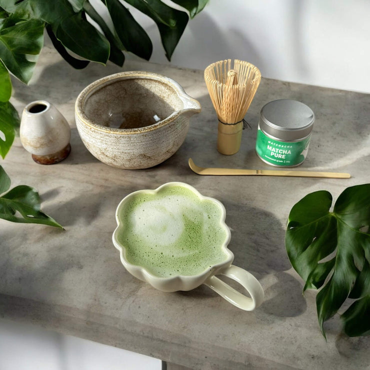 Matcha-sæt med alt nødvendigt til matcha-tilberedning, inklusive bambuspiskeris og skål – ved siden af en skummende, cremet matcha latte.